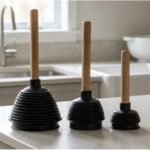 Kit com três desentupidores sanfonados de borracha com cabo, em tamanhos diferentes, apoiados sobre bancada de cozinha